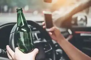 A person holds a beer bottle in one hand and a smartphone in the other while sitting behind the steering wheel of a car.