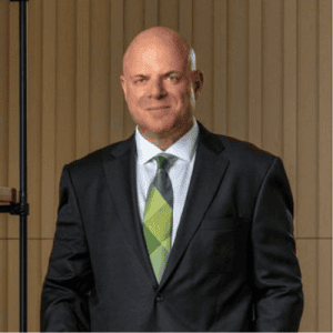 A man in a dark suit and green tie, reminiscent of a Philadelphia Car Accident Lawyer, stands in front of a striped wall, smiling slightly.