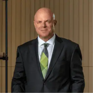 A man in a dark suit and green tie, reminiscent of a Philadelphia Car Accident Lawyer, stands in front of a striped wall, smiling slightly.