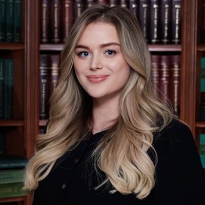 A person with long, wavy blond hair is standing in front of bookshelves filled with books and smiling slightly.