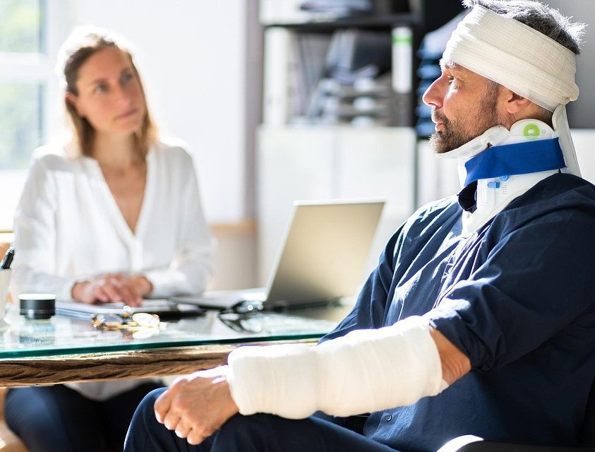 A man with bandages and a neck brace sits in a wheelchair facing a woman at a desk. The woman appears to be listening attentively.