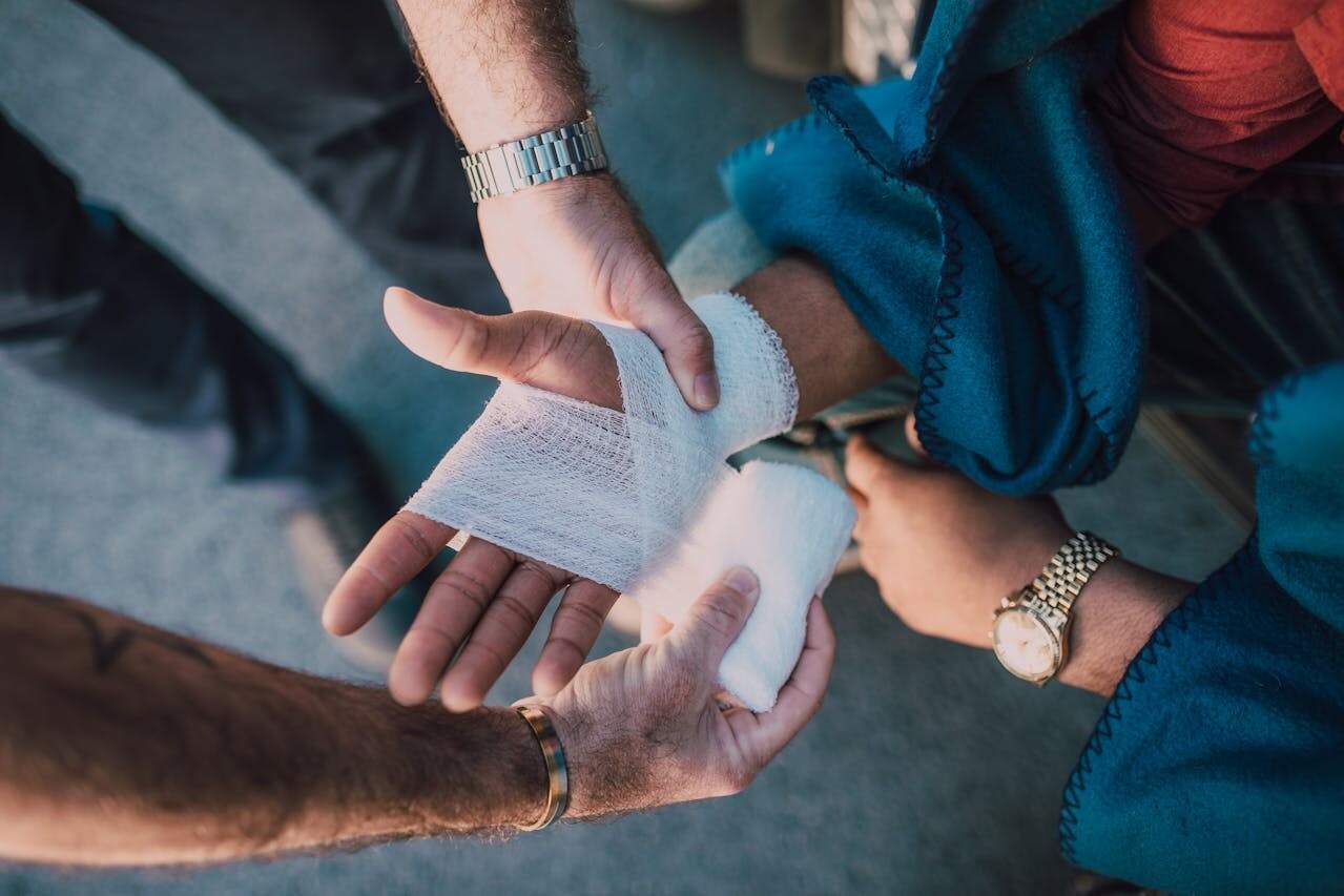 A person wraps another person's injured hand with a white bandage. Both individuals are wearing wristwatches.
