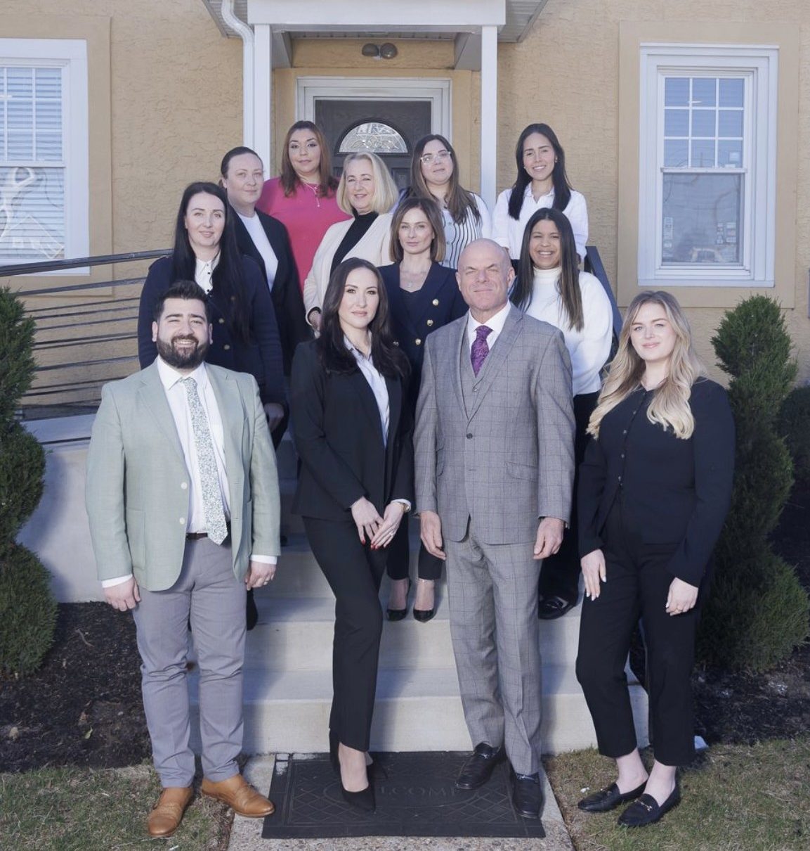 A group of twelve professionally dressed people pose for a photo on the steps outside a tan building with white trim.