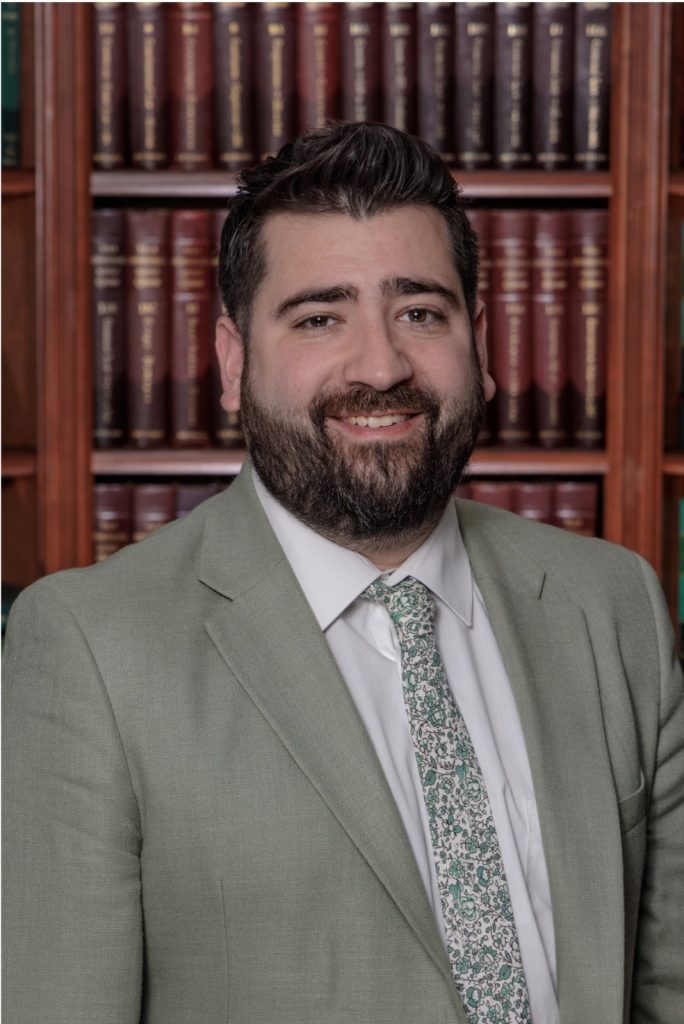 A man with dark hair and a beard wearing a light gray suit and patterned tie stands in front of bookshelves filled with books.