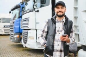 smiling truck driver holding smartphone standing in front of commercial trucks
