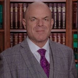 A bald man in a gray plaid suit with a purple tie is standing in front of bookshelves filled with legal books. A bald man in a gray plaid suit with a purple tie is standing in front of bookshelves filled with legal books.
