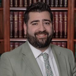 A man with dark hair and a beard, wearing a light gray suit, white shirt, and patterned tie, poses in front of bookshelves filled with books. A man with dark hair and a beard, wearing a light gray suit, white shirt, and patterned tie, poses in front of bookshelves filled with books.