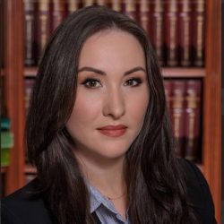 A woman with long brown hair wearing a blazer and striped shirt poses in front of bookshelves filled with legal books. A woman with long brown hair wearing a blazer and striped shirt poses in front of bookshelves filled with legal books.