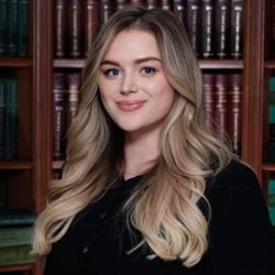 A person with long, wavy blond hair is standing in front of bookshelves filled with books and smiling slightly. A person with long, wavy blond hair is standing in front of bookshelves filled with books and smiling slightly.